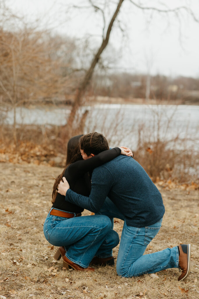 Man proposing to woman during a surprise proposal in Iowa, embracing on one knee near the river in Cedar Falls with winter trees in the background.