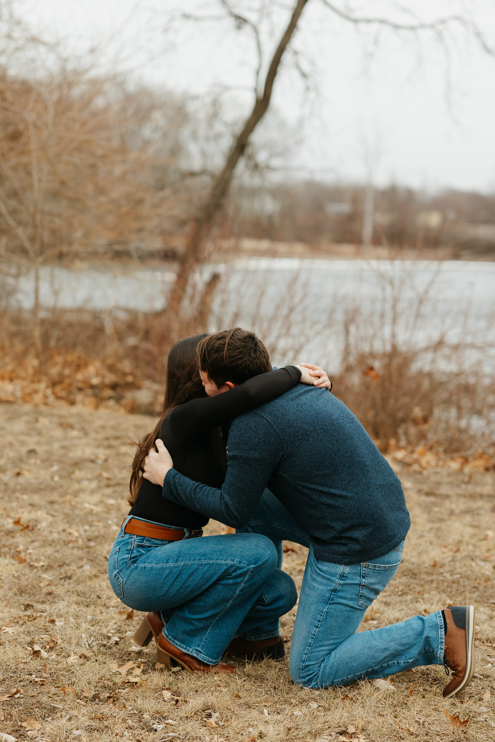 Man proposing to woman during a surprise proposal in Iowa, embracing on one knee near the river in Cedar Falls with winter trees in the background.